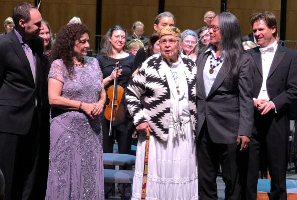 Following the 2018 performance of “How Can You Own the Sky?” in Grants Pass, tribal elder Agnes Emma Baker Pilgrim (center, in white dress) asked to be taken onstage, where she blessed the audience. She was joined by (from left) composer Ethan Gans-Morse, lyricist Tiziana DellaRovere, Native drummer and singer Brent Florendo, and conductor Martin Majkut of the Rogue Valley Symphony. Photo courtesy: Ethan Gans-Morse